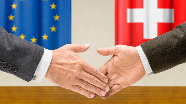Deux mains se serrent la main. En arrière-plan, un drapeau de l'UE et un drapeau suisse